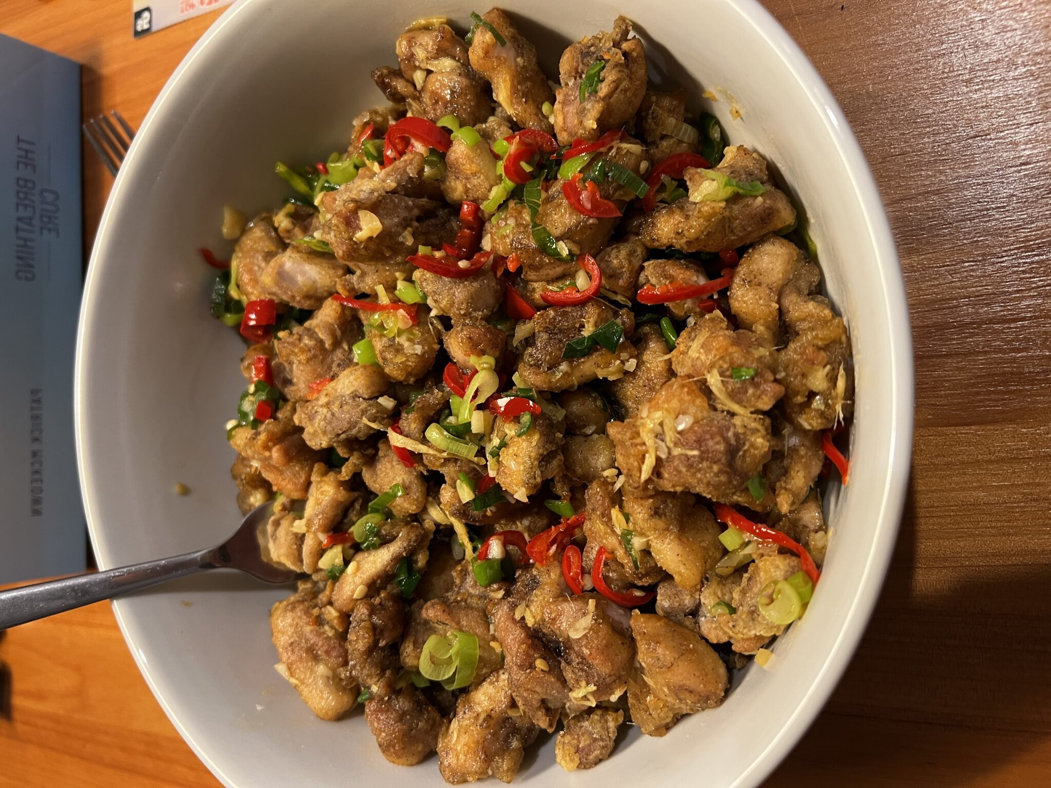 A white bowl of crispy salt and pepper chicken with sliced red chilli and spring onions on a wooden table